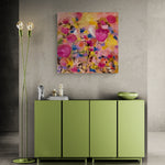 Load image into Gallery viewer, A square abstract floral wall art print with vibrant pink, yellow, and blue brushstrokes hangs on a textured gray wall above a green sideboard, with a matching green floor lamp on the left.
