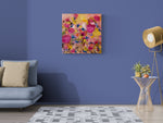 Load image into Gallery viewer, A square abstract floral wall art print with vibrant pink, yellow, and blue brushstrokes, displayed on a blue wall in a modern living room setting with a gray sofa, side table, and gold floor lamp.
