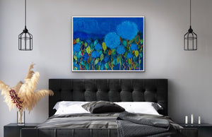 Predominantly blue floral painting shown  in a bedroom setting above e a charcoal grey headboard and linens.