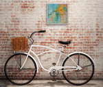Load image into Gallery viewer, This original abstract flower painting in gold, teal, and green is sweet and simultaneously quirky with a vintage vibe and a feeling of nostalgia. It has a glossy finish. Displayed on a brick wall above a bicycle.
